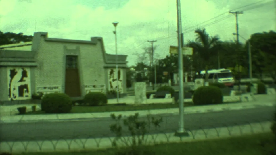 1973 Yucatan 8mm frame showing the low mural-sided building, roadside plantings, a lamp post, parked cars, and palm-like trees.