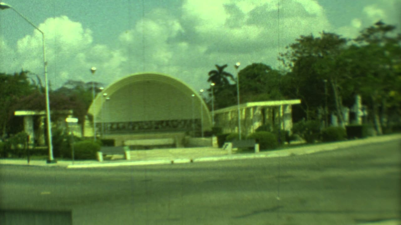 Yellow domed building beside a quiet paved road with shrubs, trees, streetlights, and clouds.