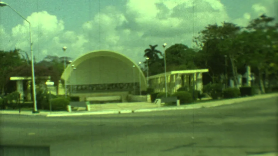 1973 Yucatan view of a yellow domed building across a paved street, with landscaping, trees, streetlights, and a cloudy sky.