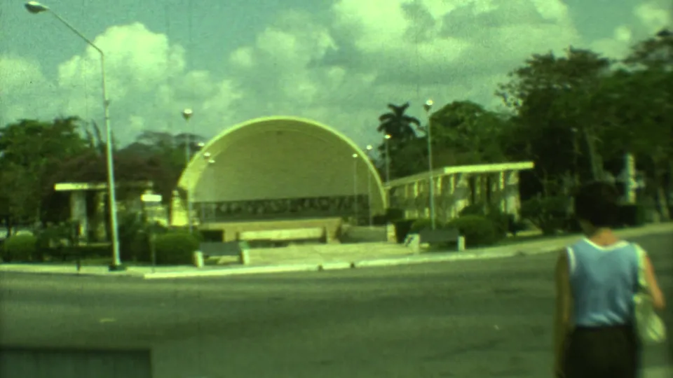1973 Yucatan footage shows a woman in a blue sleeveless top and dark skirt walking in the foreground before a yellow domed building.