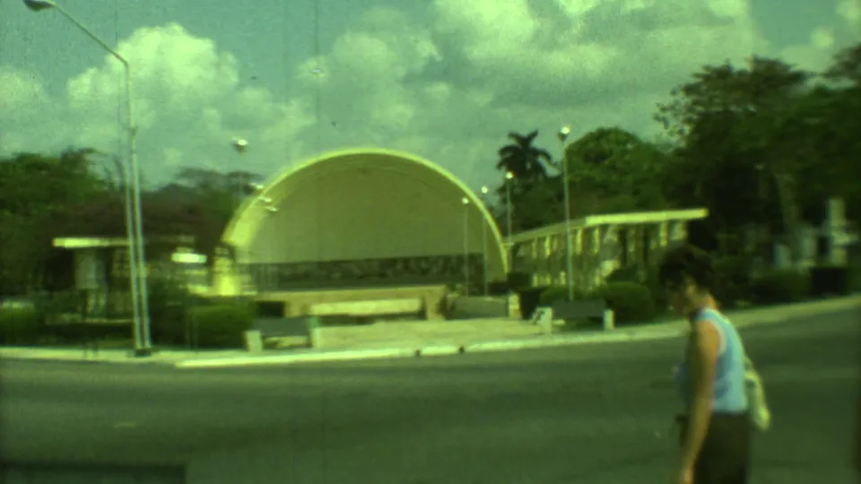 1973 Yucatan park view with arched pavilion structures, benches, palms, and a pedestrian in casual clothing near the road.