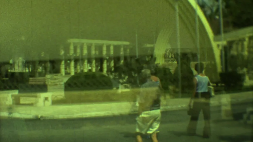 Green-shifted 8mm frame showing pedestrians in casual 1970s clothing near benches and an arched park pavilion.