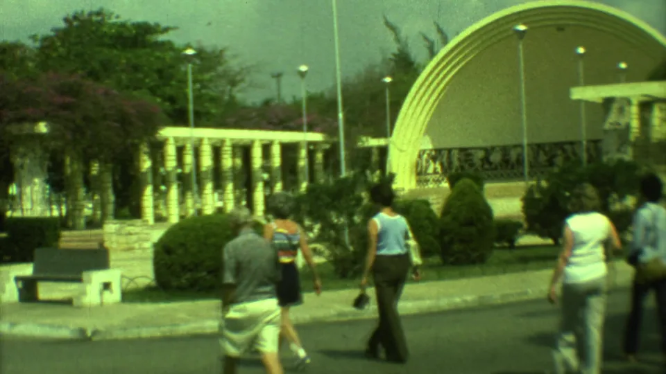 1973 Yucatan park scene with multiple pedestrians, shrubs, benches, lamp posts, and an arched pavilion in the background.