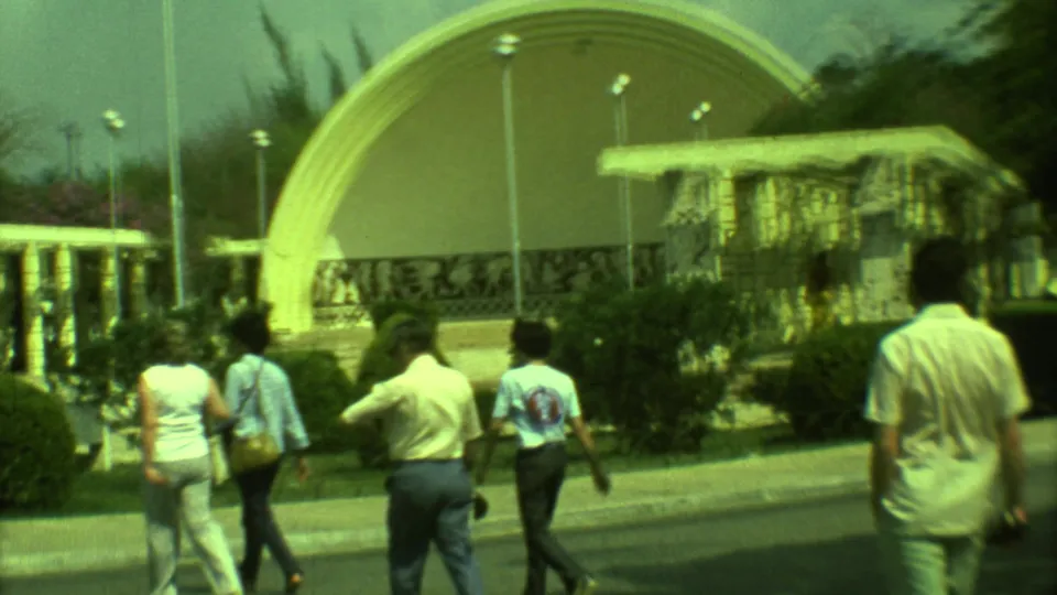 1973 Yucatan 8mm frame of visitors walking past shrubs and lampposts toward a large curved outdoor exhibit structure.