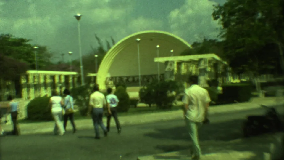 Wide 1973 Yucatan home-movie view of pedestrians walking near a curved dome structure in a landscaped public outdoor area.