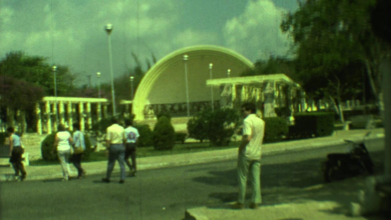 Visitors walk near an arched pavilion and columns in a sunny public space.