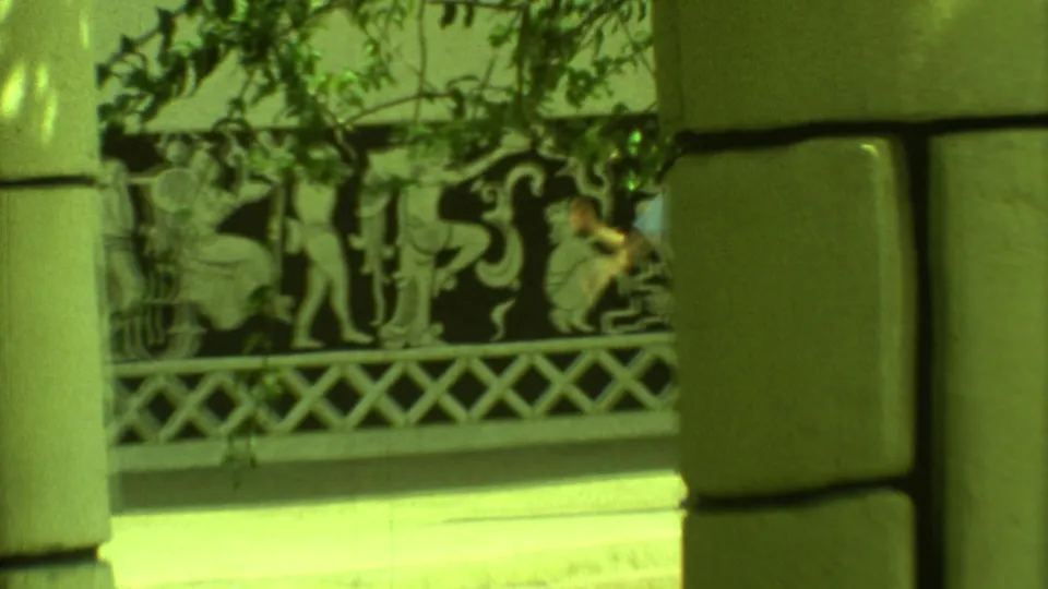 1973 Yucatan 8mm frame showing visitors near Mayan-style wall panels, partly hidden by large stone columns and foliage.