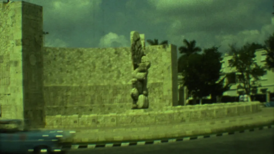 1973 Yucatan 8mm wide shot showing the monument beside a stone wall, road edge, trees, and background buildings.