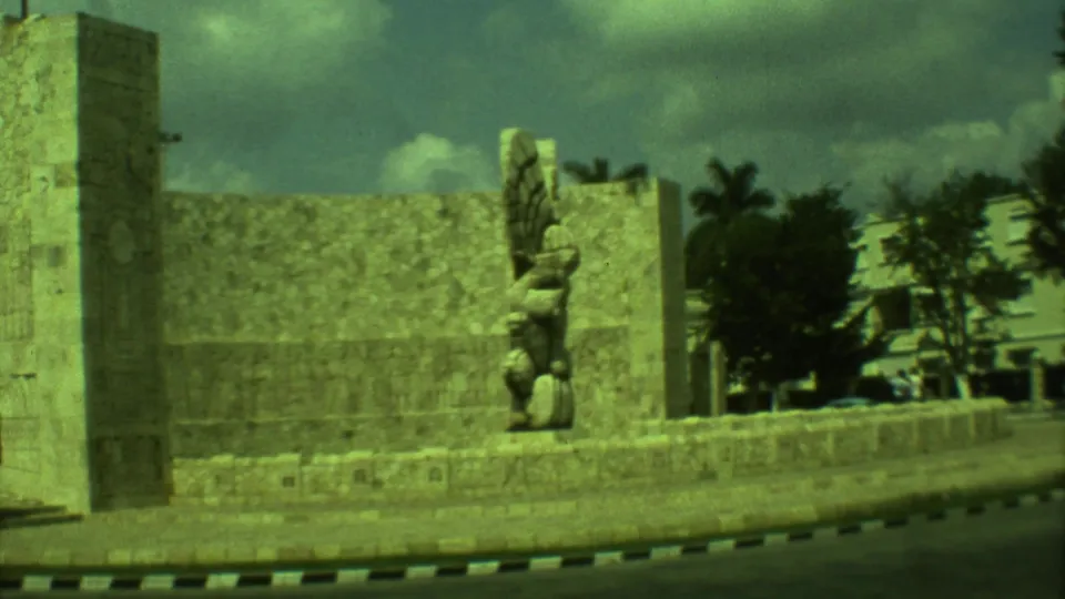 1973 Yucatan home-movie frame showing the carved stone figure, wall, pillar, trees, road edge, and distant buildings.