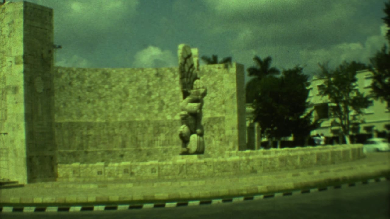 Stone monument beside a road with carved slabs, a winged sculpture, palm trees, and cloudy sky.