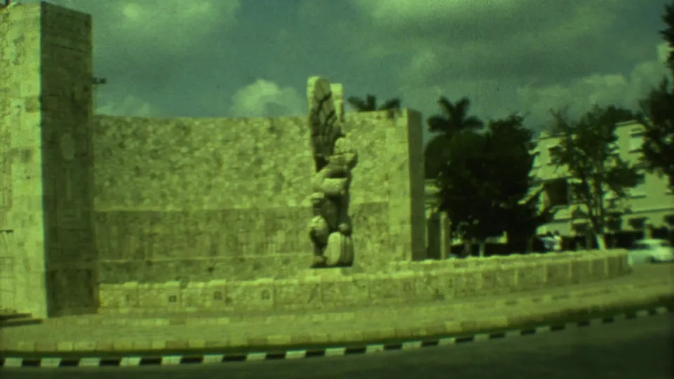 1973 8mm view in Yucatán, Mexico, showing a Mayan-style stone monument at a roundabout with a winged sculpture and roadside curb.