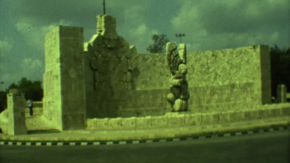 1973 Yucatán street view with a Mayan-style stone monument, winged sculpture, curb, and a distant person near the roadside.