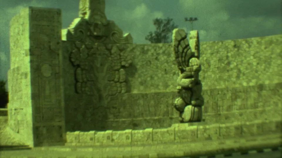Final 1973 8mm frame of the Yucatán roundabout monument, showing carved stone surfaces, a winged sculpture, road curb, and trees.