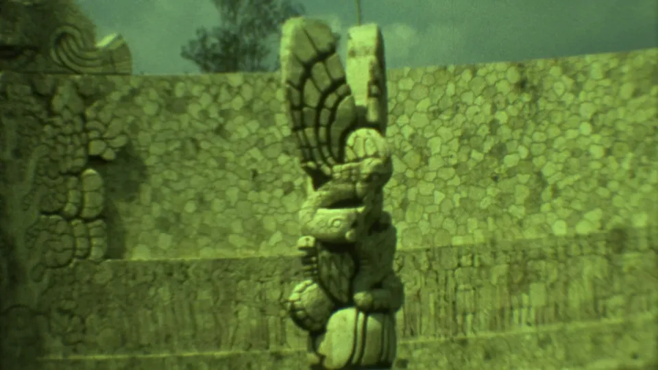 1973 Yucatan home-movie frame with a tall sculptural stone form, weathered wall, and dry vegetation.