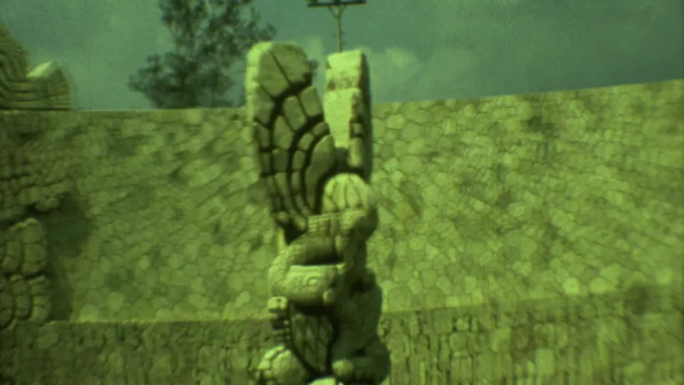 1973 Yucatan 8mm frame showing a weathered Maya-style stone sculpture near a high stone wall and cloudy sky.