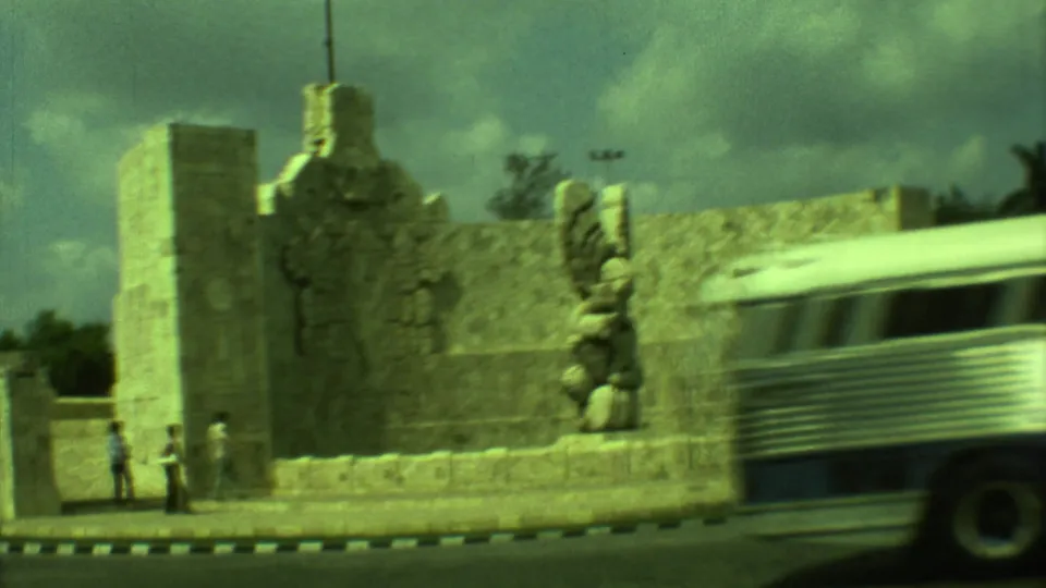 Wide 8mm view of a Yucatan stone monument as a bus passes and small pedestrians stand near the curb.