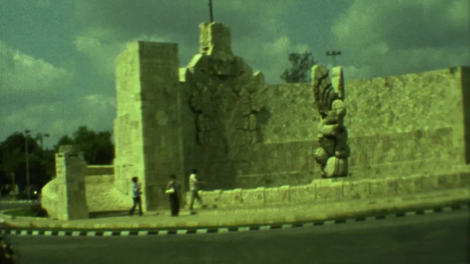 1973 Yucatan frame showing pedestrians near a stone sculpture and wall at a historic roadside site.