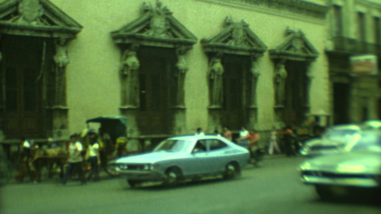 Classic cars pass pedestrians outside a colonial building with ornate window surrounds.