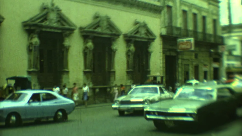 A light blue classic car and motion-blurred traffic move past pedestrians and colonial building windows in 1973 Yucatan footage.