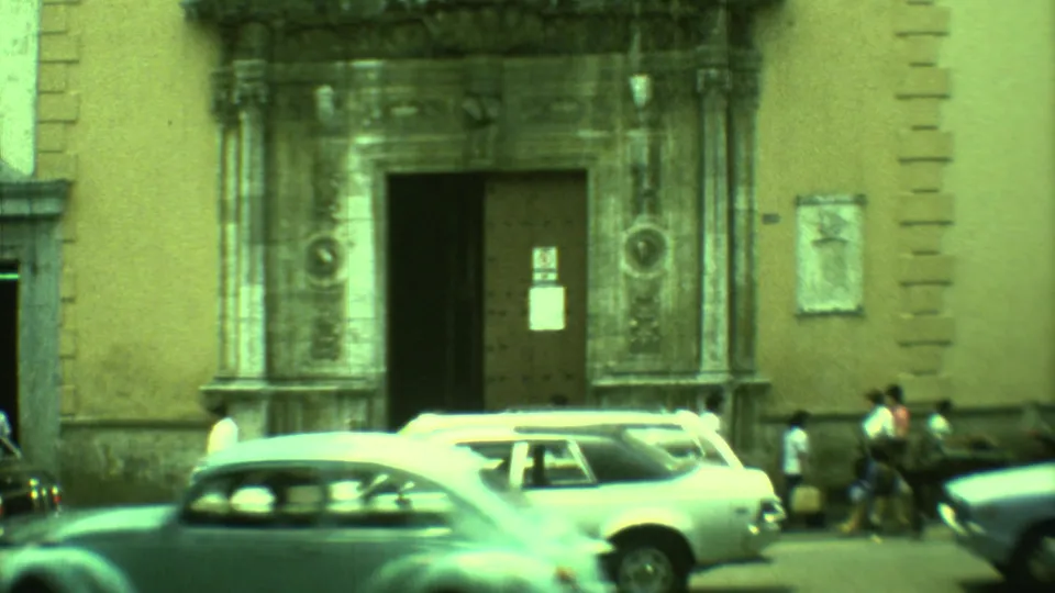 Classic cars, including a light-colored wagon, pass a large ornate doorway on a Yucatan street in 1973 home-movie footage.