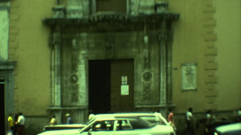 Pedestrians stand near cars below a large colonial doorway and balcony in 1973 Yucatan street footage.