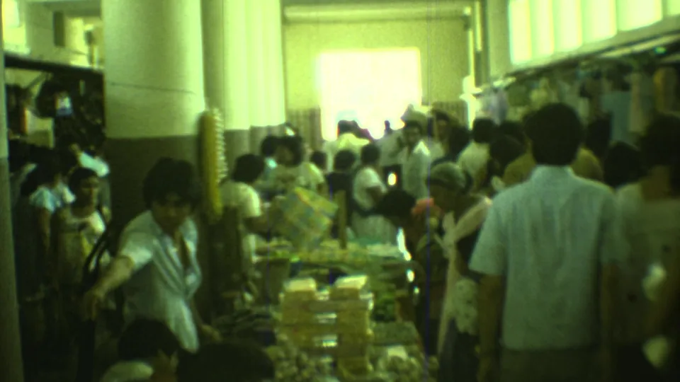 1973 Yucatan home-movie frame showing adults in a busy indoor market aisle with tables, goods, and bright windows.