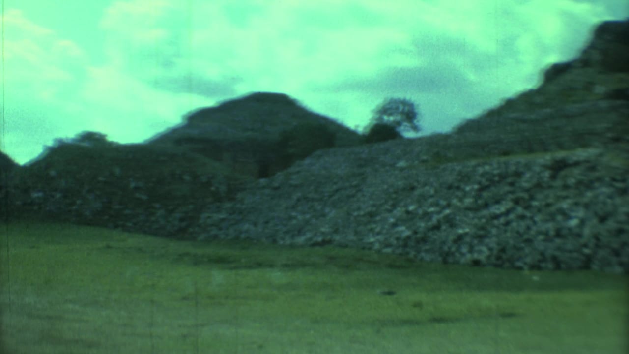 Stone ruin wall and rounded mound seen across grass under green tint.