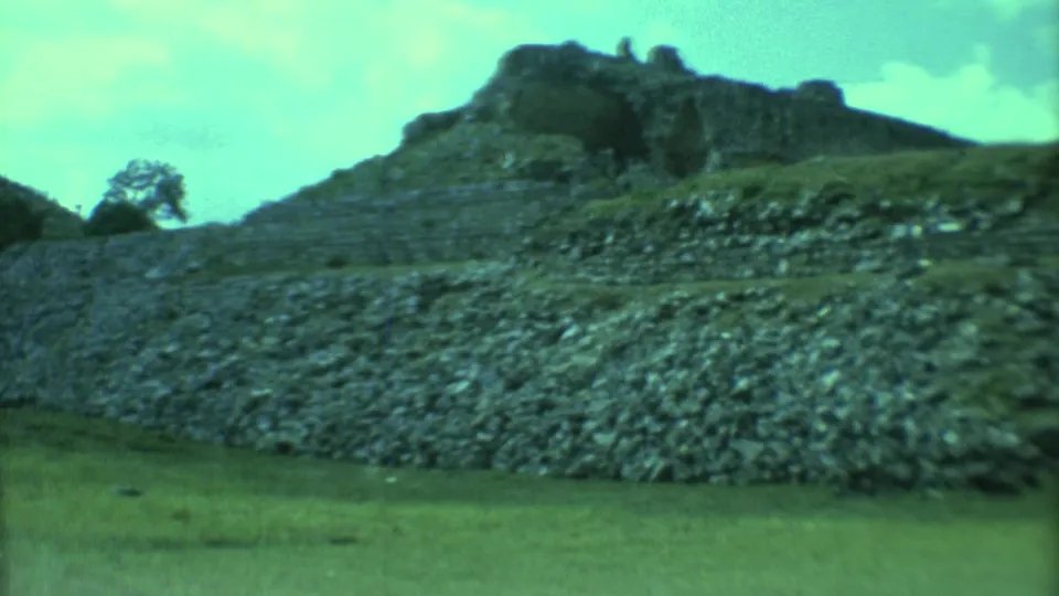 1973 Yucatan home-movie frame with a raised rocky ruin structure, stone wall, grass, and cloudy sky in aged 8mm color.