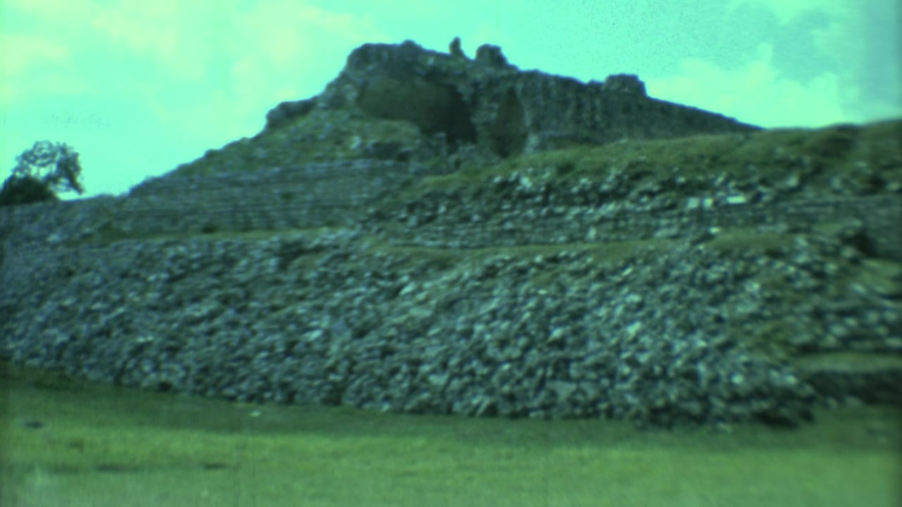 Stone pyramid and stepped platform rising above a grassy plain under a pale sky.