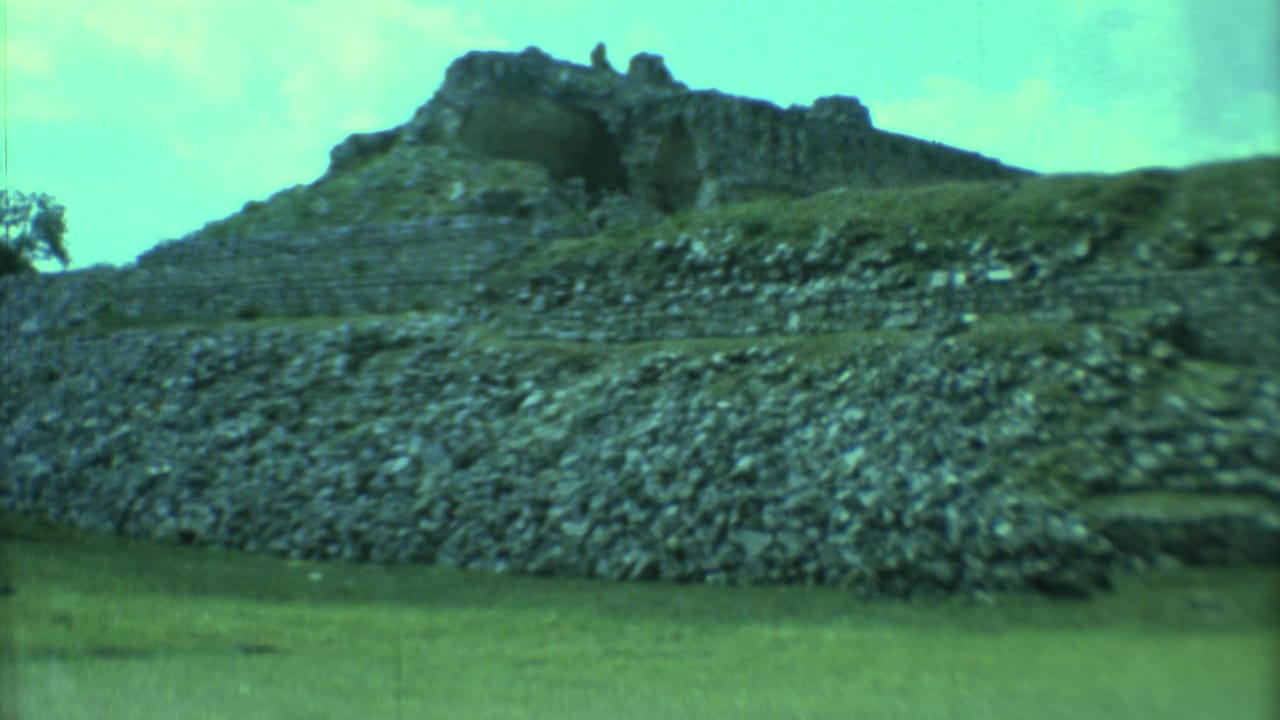 Rocky stepped ruin and low pyramid form seen across a grassy foreground.