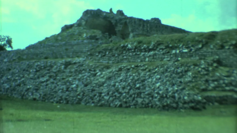 1973 Yucatan home-movie frame showing a rocky stepped ruin and low pyramid profile across an open grassy plain.