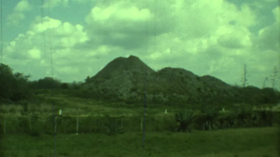 1973 8mm Yucatan frame with low stone pyramids or mounds, scrub vegetation, grass, and a partly cloudy sky.