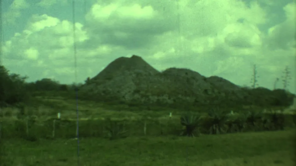 Vintage 1973 Yucatan footage frame showing a low stone pyramid, rounded mound, grass, scrub plants, and a partly cloudy sky.