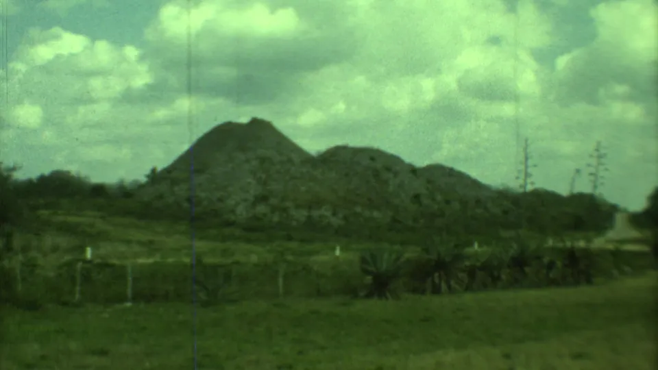 1973 8mm Yucatan view of rocky mounds, grass, fence posts, sparse plants, and a cloudy sky with a green film tint.