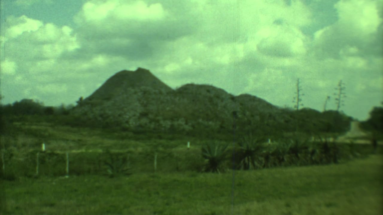 1973 8mm Yucatan landscape with rock piles, grass, a narrow dirt path, fence posts, and distant sparse vegetation.