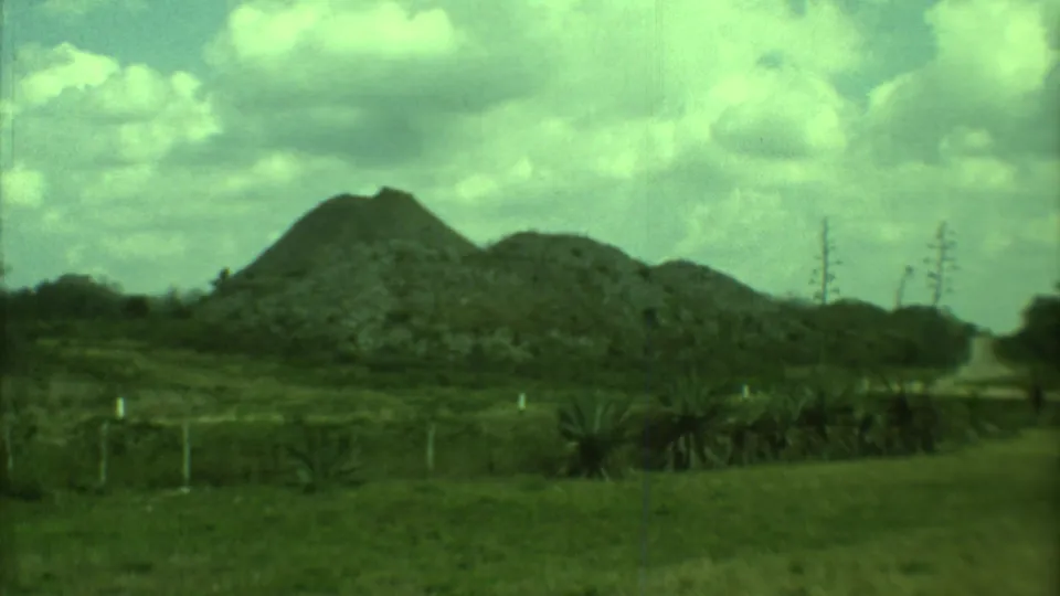 1973 8mm Yucatan landscape with rock piles, grass, a narrow dirt path, fence posts, and distant sparse vegetation.