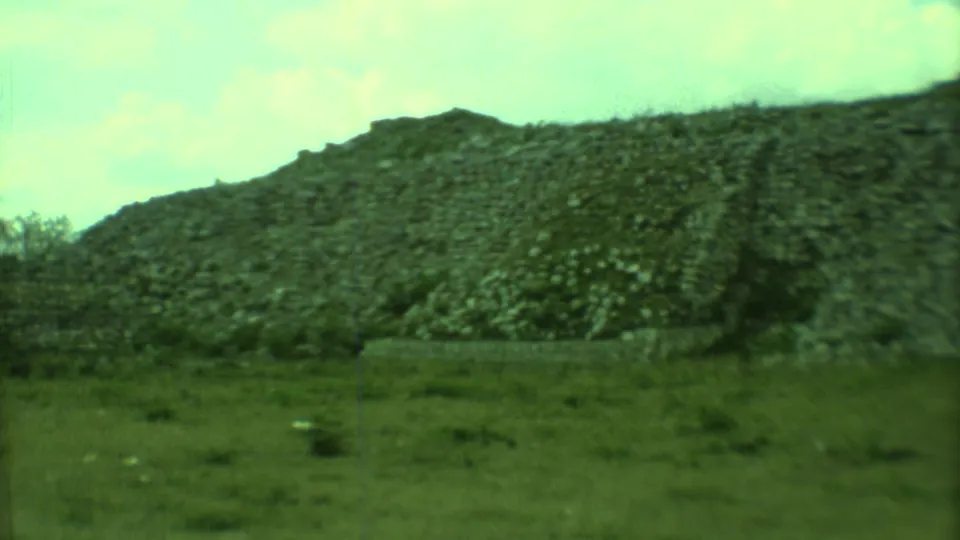 Closer 1973 Yucatan 8mm view of a rocky slope, low grass, scattered bushes, and pale sky, with film grain and soft focus.