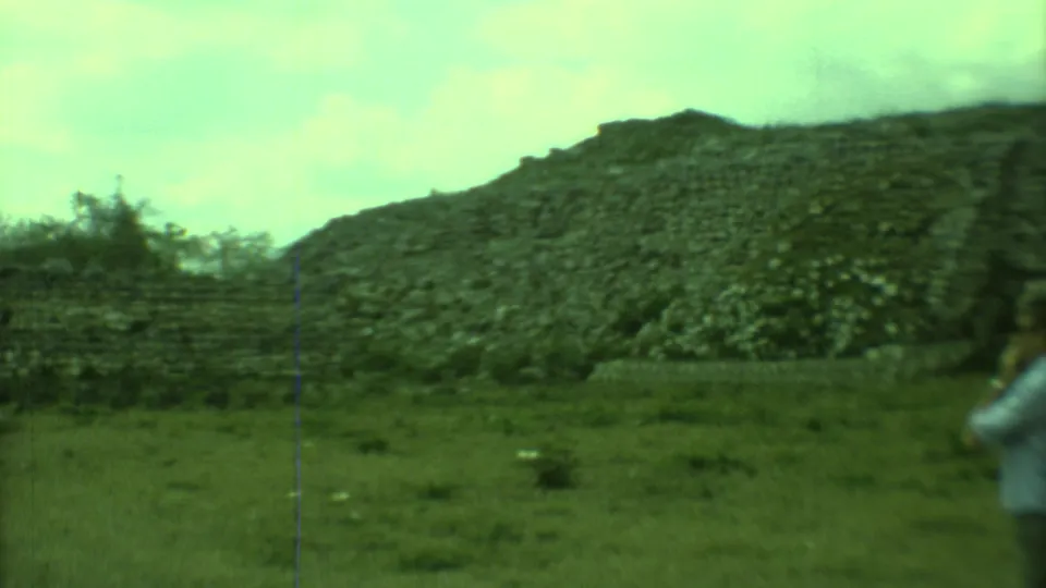 1973 8mm Yucatan frame showing a rocky hillside and grassy foreground, with a man in a light shirt partly visible at the right edge.