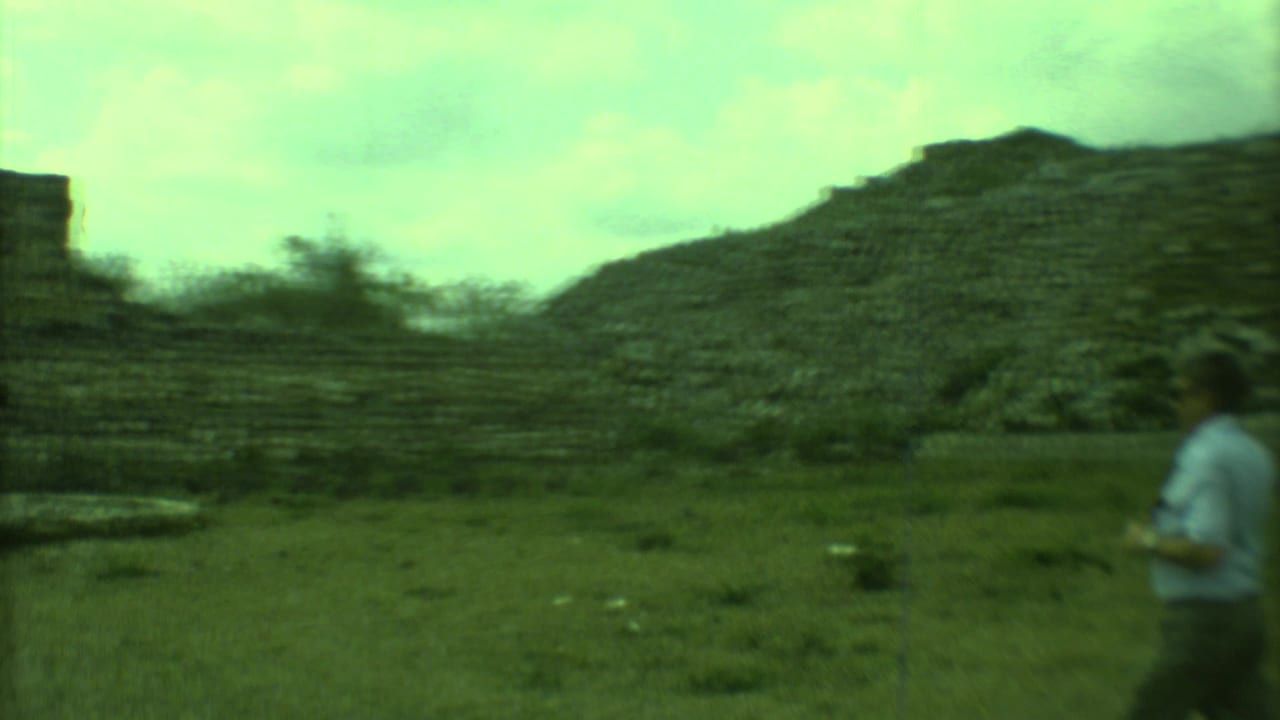 Tourists walking near stepped stone ruin structures under a cloudy sky.