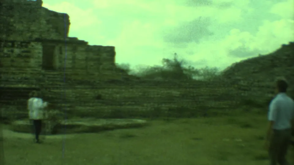 Tourists in casual 1970s clothing move near a weathered stone ruin building with dark window openings.