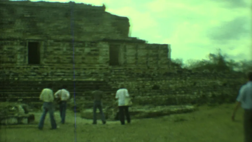 A group of adult visitors crosses the grassy foreground below a stepped stone ruin facade in Yucatan, Mexico.