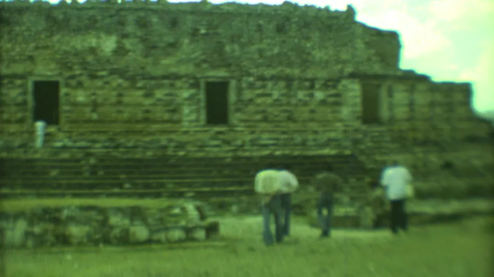 Adults stand near a low stone platform with a larger weathered ruin wall and dark openings behind them.