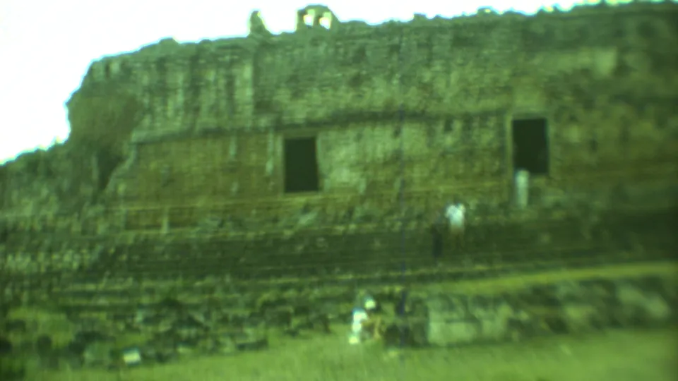Green-tinted 8mm view of a Mayan ruin facade in Yucatan with a visitor standing near the stepped platform.