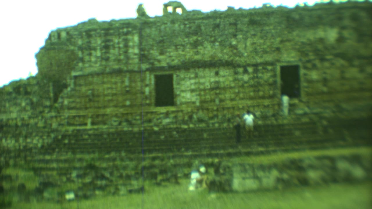 Tourists near a stepped stone ruin with dark doorways in bright sun.