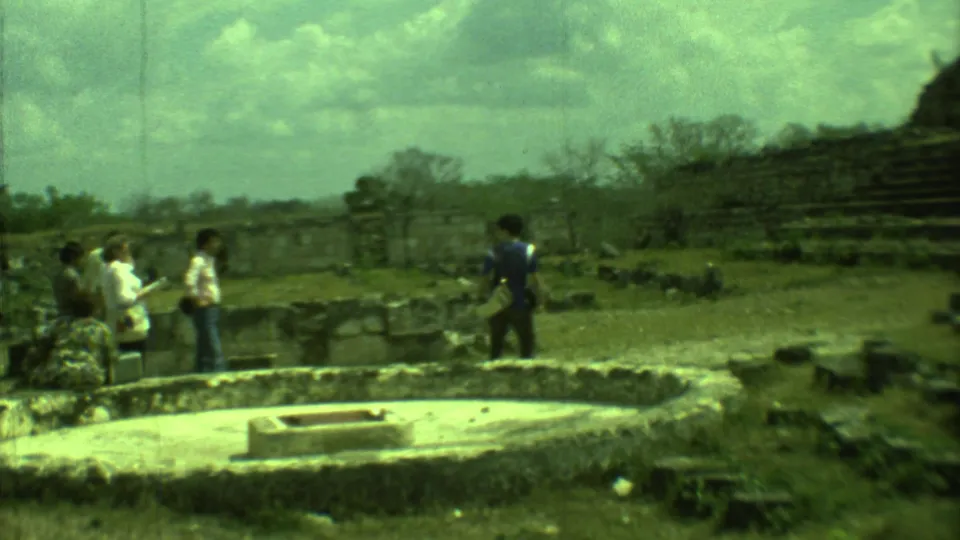 1973 Yucatan home-movie frame of visitors looking across low stone ruins and a circular stone feature.