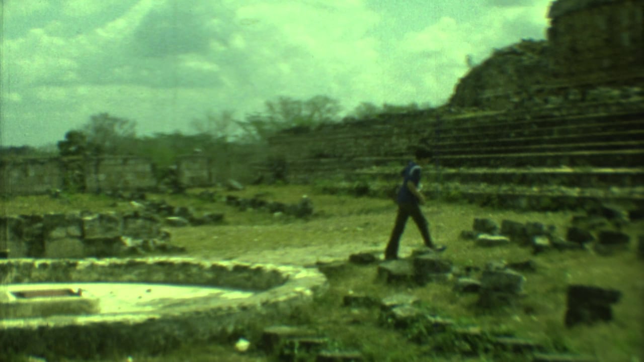 A man walks past low stone remains with a stepped ruin rising behind him.