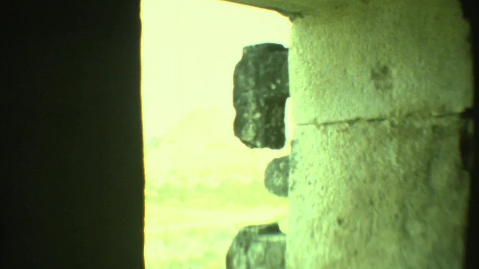 From inside or beside a stone structure, the 8mm frame shows a dark doorway edge, rough masonry, and bright ruin landscape outside.