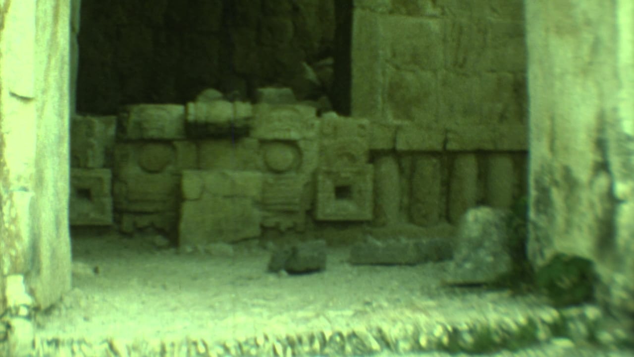 A shadowed Mayan ruin chamber in Yucatan appears through a weathered doorway, with carved stone pieces along the back wall.