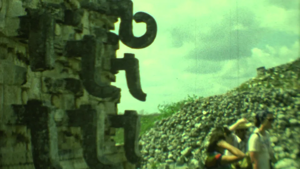Tourists in casual 1970s clothing stand near carved stone architecture at a Yucatan ruin under a cloudy sky.