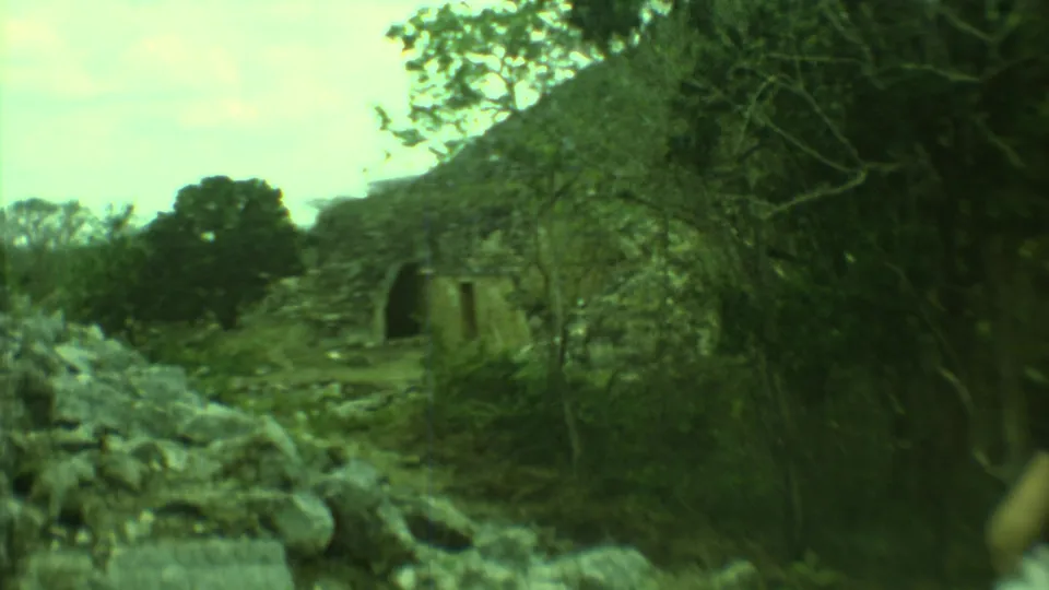 Handheld 8mm footage looks past dense branches and rubble toward a small arched stone ruin at the Yucatan site.